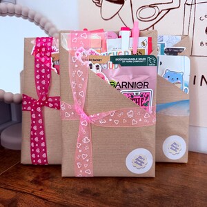 May include: Three rectangular brown paper gift envelopes tied with decorative ribbons. The center envelope features a pink ribbon with white heart outlines and contains a Garnier face mask sachet, a pink pen, and various stickers. Each envelope has a small round sticker.