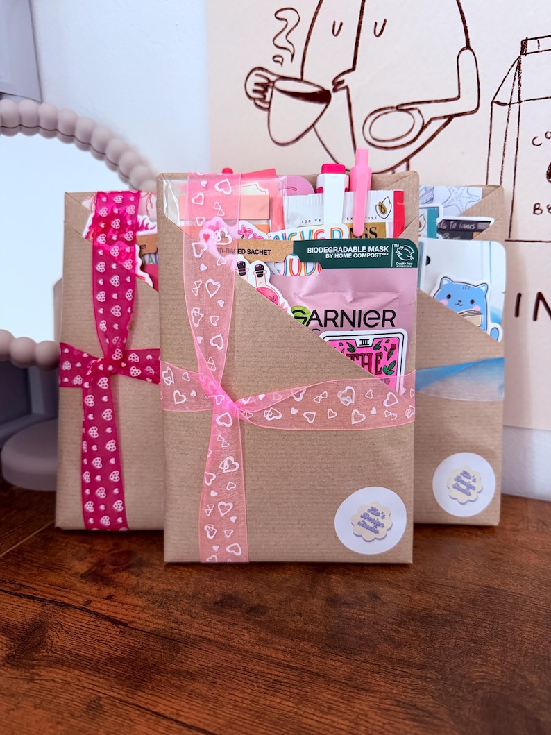 May include: Three rectangular brown paper gift envelopes tied with decorative ribbons. The center envelope features a pink ribbon with white heart outlines and contains a Garnier face mask sachet, a pink pen, and various stickers. Each envelope has a small round sticker.