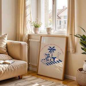 May include: A framed art print with the words "BOOKS, BREEZE, AND BAREFOOT DREAMS" depicts a woman in a swimsuit reading on a beach towel under a palm tree. The print is in a light wood frame, near a window with a beige sofa and a potted plant.
