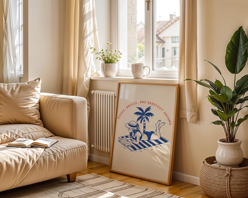 May include: A framed art print with the words "BOOKS, BREEZE, AND BAREFOOT DREAMS" depicts a woman in a swimsuit reading on a beach towel under a palm tree. The print is in a light wood frame, near a window with a beige sofa and a potted plant.