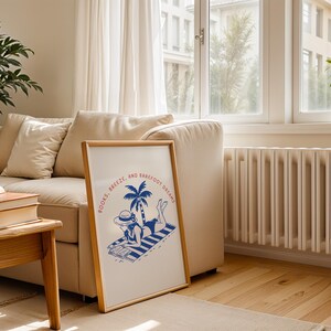 May include: A framed print with a beach scene illustration. The artwork features a person in a hat reading on a striped towel under a palm tree. The text "BOOKS, BREEZE, AND BAREFOOT DREAMS" is printed in red and blue.