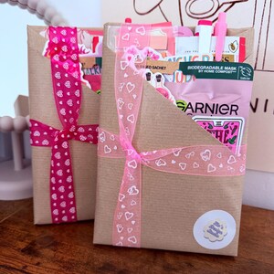 May include: Two rectangular brown paper gift envelopes tied with pink heart-patterned ribbons. The envelopes contain various items including a Garnier biodegradable mask sachet, stickers, and pens. A small circular sticker with text is on the front.