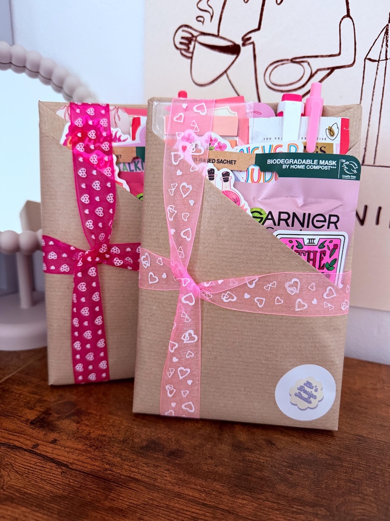 May include: Two rectangular brown paper gift envelopes tied with pink heart-patterned ribbons. The envelopes contain various items including a Garnier biodegradable mask sachet, stickers, and pens. A small circular sticker with text is on the front.