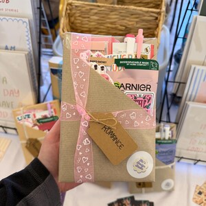 May include: A rectangular brown paper gift package wrapped with a pink ribbon featuring a white heart pattern. A brown card tag labeled ROMANCE is attached to the ribbon. The package contains a Garnier face mask, pens, and stickers.