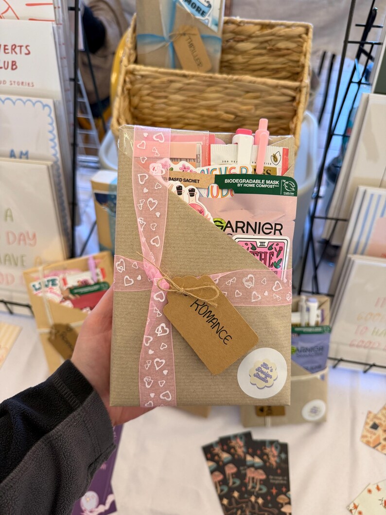 May include: A rectangular brown paper gift package wrapped with a pink ribbon featuring a white heart pattern. A brown card tag labeled ROMANCE is attached to the ribbon. The package contains a Garnier face mask, pens, and stickers.
