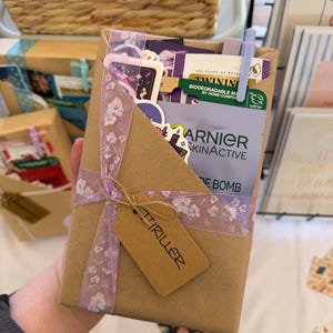 May include: A rectangular brown paper gift package containing various items including a Garnier SkinActive sheet mask, a Twinings tea bag, and stickers. The package is wrapped with a light purple floral ribbon and features a brown tag labeled THRILLER.