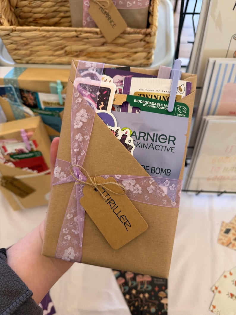 May include: A rectangular brown paper gift package containing various items including a Garnier SkinActive sheet mask, a Twinings tea bag, and stickers. The package is wrapped with a light purple floral ribbon and features a brown tag labeled THRILLER.