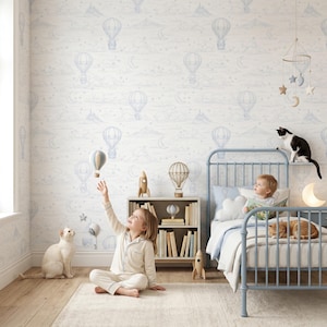 Op de afbeelding: Behang met een herhalend patroon van lichtblauwe heteluchtballonnen, maansikkels en sterren op een witte achtergrond. Het ontwerp bevat lijntekeningen van wolken en bergsilhouetten, wat zorgt voor een hemelse esthetiek.