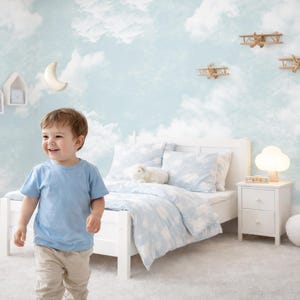 May include: A child's bedroom with a light blue and white cloud-themed wallpaper. The room features a white bed with cloud-patterned bedding, wooden airplanes, and a white bedside table with a cloud-shaped lamp. A young child in a blue top and beige trousers smiles.
