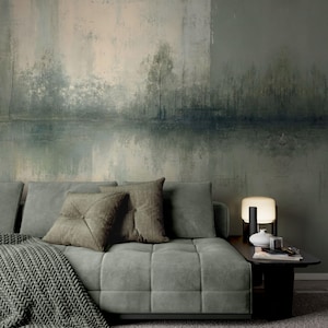 Op de afbeelding: Een moderne woonkamer met een lichtgroene, vierkant gestikte bank, olijfkleurige kussens en een gebreide deken. De achtergrond is een groot, abstract landschapsschilderij in groen- en grijstinten, met een bijzettafel en lamp.