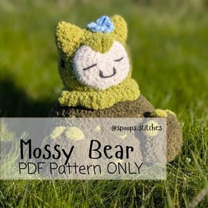 Puede incluir: Un oso de ganchillo, llamado "Mossy Bear", con cuerpo verde claro, cara blanca y una pequeña flor azul. El oso tiene los ojos cerrados y una boca sonriente. El texto "Mossy Bear PDF Pattern ONLY" se muestra en la imagen.