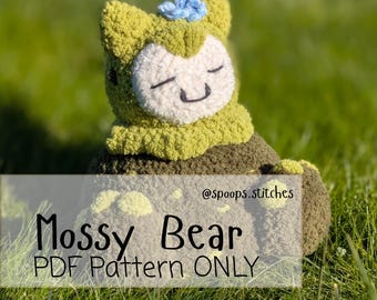 May include: A crocheted Mossy Bear, with a light green body, a white face, and a small blue flower. The bear has closed eyes and a smiling mouth. The text "Mossy Bear PDF Pattern ONLY" is displayed on the image.