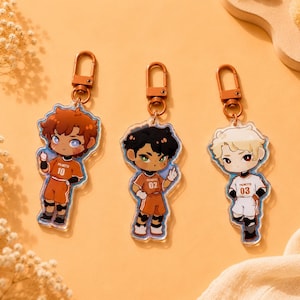Peut inclure: Trois porte-clés en acrylique représentant des personnages de style chibi en tenue de sport. Le personnage de gauche a les cheveux bruns et porte le numéro 10 orange, le centre a les cheveux noirs et le numéro 02 orange, et la droite a les cheveux blonds et le numéro 03 blanc.