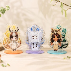 以下が含まれることがあります： 詳細なデザインの、アニメスタイルのフィギュアが特徴の3つのアクリル製キャラクタースタンド。フィギュアは、角や花のヘッドピースなど、ユニークな衣装とアクセサリーを身に着けています。各スタンドは、オレンジ、紫、ティール色のカラフルなベースを持っています。