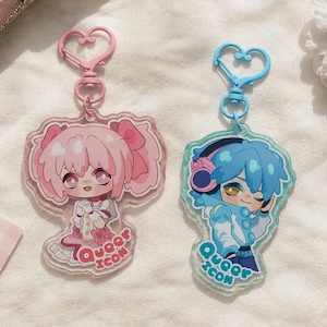 Peut inclure: Deux porte-clés en acrylique avec des personnages de style anime. L'un présente un personnage rose avec un fermoir en forme de cœur rose, l'autre un personnage bleu avec un fermoir en forme de cœur bleu. Les deux porte-clés ont le texte "Queer Icon".