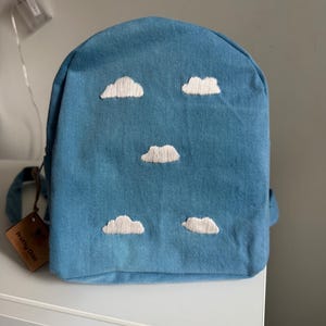 May include: A blue denim backpack featuring five embroidered white clouds on the front panel. A brown rectangular tag attached to the side strap displays the text Pretty Odd in black lettering. The backpack has a rounded top and a structured shape.