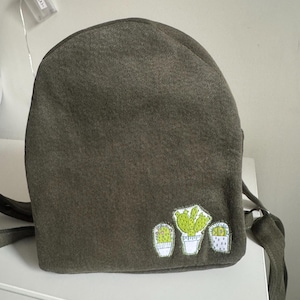 May include: An olive green backpack made of a textured fabric featuring an embroidered patch at the bottom right corner. The patch displays three potted cacti in shades of light green and white with visible stitching details.