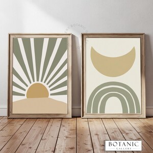 May include: Two framed abstract art prints. One features a sunburst design in olive green, tan, and cream. The other has a crescent moon and rainbow in similar colours. Both are in wooden frames, with the "BOTANIC GALLERY" logo at the bottom.
