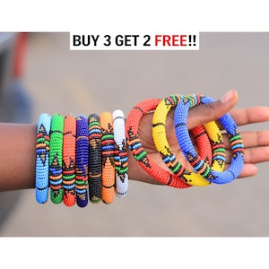 May include: A collection of round beaded bracelets featuring vibrant multi-colored patterns in shades of blue, green, orange, red, yellow, black, and white. The text BUY 3 GET 2 FREE!! is displayed at the top of the image above the bracelets.