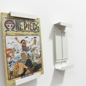 Può includere: Una mensola bianca a parete che tiene un fumetto manga. La copertina del fumetto presenta illustrazioni colorate e il titolo "ONE PIECE". Una seconda mensola simile è montata a destra.