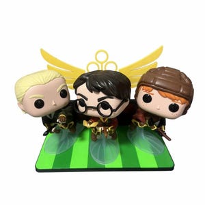 Può includere: Tre personaggi Funko Pop di una serie fantasy. Le figure sono su una base a strisce verdi e bianche con un emblema giallo. I personaggi indossano uniformi e tengono scope.
