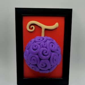 Puede incluir: Una obra de arte enmarcada que presenta una fruta morada con remolinos y un tallo beige sobre un fondo rojo. La fruta es el punto focal, encerrada en un marco negro. El diseño es una representación 3D de una fruta estilizada.