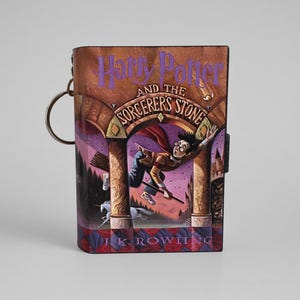 Peut inclure: Un portefeuille rectangulaire arborant la couverture de Harry Potter à l'école des sorciers de J.K. Rowling. Le motif représente un personnage volant sur un balai sous une arche, dans des tons violets et bruns, avec un anneau métallique sur le côté.