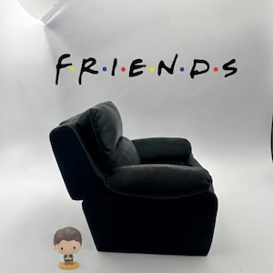 May include: A black armchair with a corduroy texture, positioned in front of a white wall. The word "FRIENDS" is written across the wall in a colorful font. A small figurine of a person is in front of the chair.