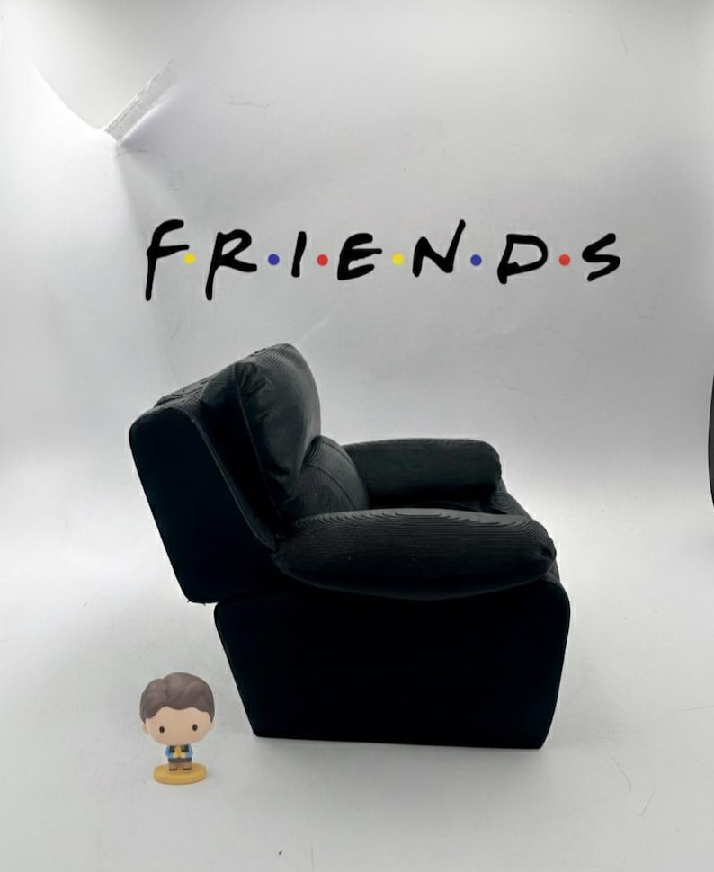 May include: A black armchair with a corduroy texture, positioned in front of a white wall. The word "FRIENDS" is written across the wall in a colorful font. A small figurine of a person is in front of the chair.
