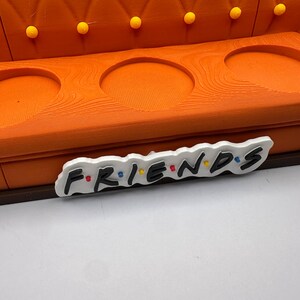 May include: An orange decorative sofa with three circular cutouts and yellow button tufting. The word "FRIENDS" is displayed in a colorful, stylized font on a white background, placed in front of the sofa.