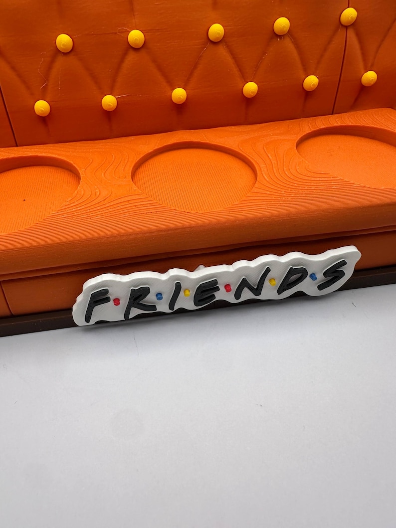 May include: An orange decorative sofa with three circular cutouts and yellow button tufting. The word "FRIENDS" is displayed in a colorful, stylized font on a white background, placed in front of the sofa.