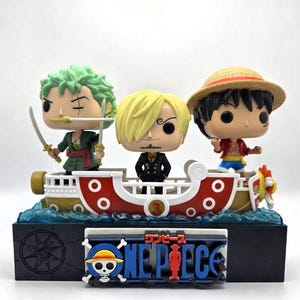 Può includere: Un oggetto da collezione di One Piece con tre personaggi su una nave, con il logo One Piece sulla base. Le figure includono un personaggio dai capelli verdi con spade, un personaggio biondo e un personaggio con un cappello di paglia.