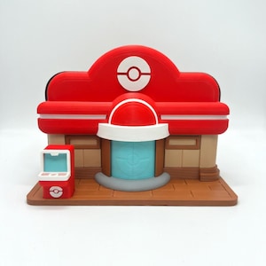 Op de afbeelding: Een 3D-geprint model van een rood en wit gebouw met een cirkelvormig logo bovenop. Het gebouw heeft een blauwe deur en een kleine rode kiosk aan de zijkant. Het model staat op een bruine basis.