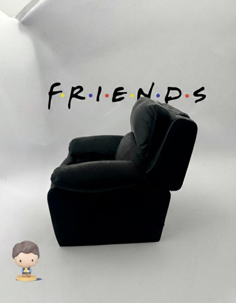 May include: A black miniature armchair with a high back and armrests. The word "FRIENDS" is written above the chair in black letters with colored dots. A small figurine of a person is in the foreground.