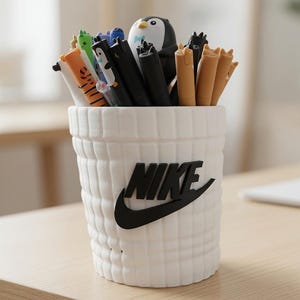 Può includere: Un portapenne cilindrico bianco con un motivo a griglia testurizzato e un logo Nike nero sul davanti. Il supporto è riempito con varie penne, incluse diverse con decorazioni a tema animale come un pinguino, gatti e altre creature.