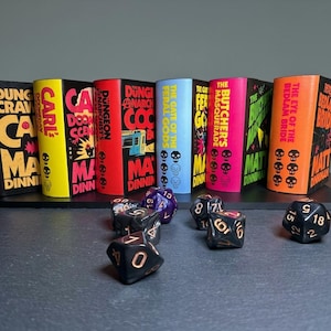 May include: Six rectangular book-shaped keychains with colourful spines featuring titles like Dungeon Crawler Carl, The Gate of the Feral Gods, and The Butcher's Masquerade by Matt Dinniman, arranged with several black polyhedral dice in the foreground.