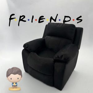 Può includere: Una poltrona reclinabile nera con il logo Friends sullo sfondo. Una piccola statuina di un uomo è di fronte alla sedia. Il logo Friends è nero con le lettere nei colori giallo, blu e rosso.