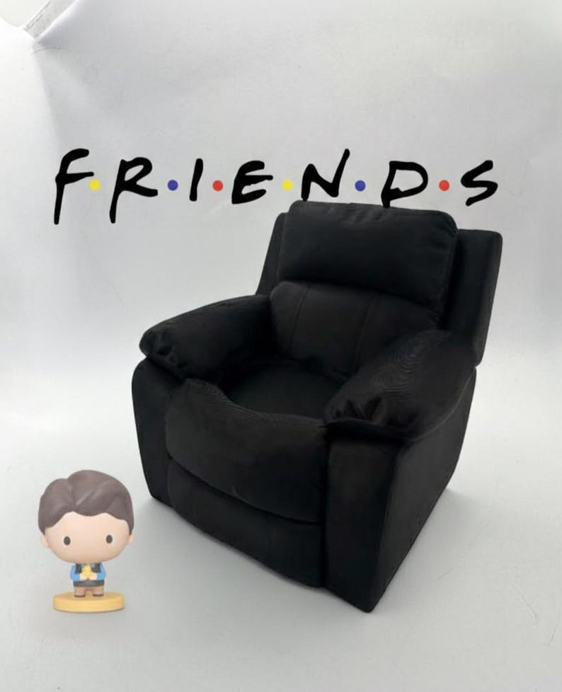 May include: A black recliner chair with a Friends logo in the background. A small figurine of a man is in front of the chair. The Friends logo is in black with the letters in the colors yellow, blue, and red.