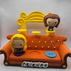 以下が含まれることがあります： 黄色いアクセントと2体のキャラクターフィギュアが付いた、詳細なオレンジ色のソファ。ソファの前面にはFriendsのロゴがあります。フィギュアは、金髪の女性と茶色の髪の女性です。
