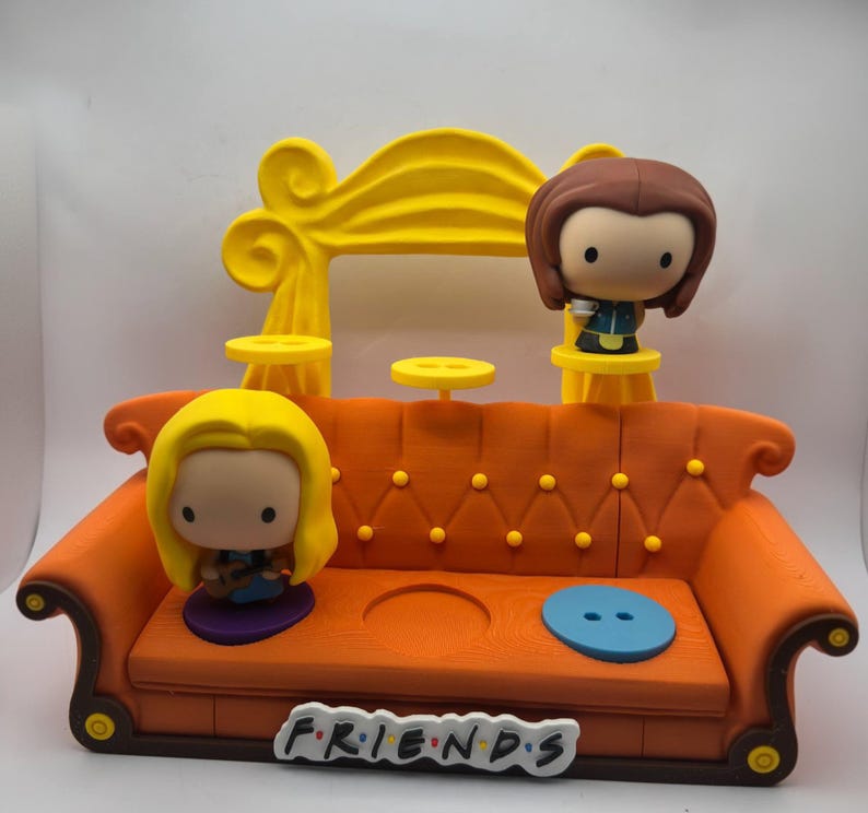 May include: A detailed orange sofa with yellow accents and two character figurines. The sofa has a Friends logo on the front. The figurines are of a woman with blonde hair and a woman with brown hair.
