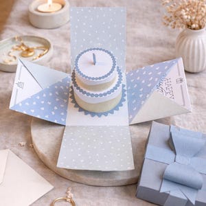 May include: A light blue pop-up birthday card with a two-tiered cake and a single candle. The card has a polka dot and heart pattern. The words "HAPPY BIRTHDAY" are printed on one panel. A small gift box with a blue ribbon is also visible.