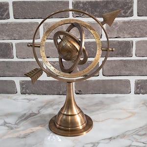 May include: A brass armillary sphere with a globe at the center, surrounded by rings and arrows. The decorative piece is mounted on a pedestal base. The background features a brick wall.