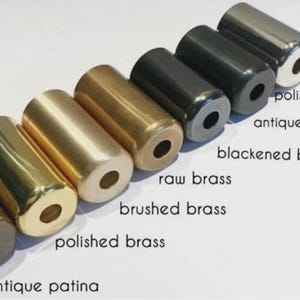 May include: A row of six cylindrical metal hardware pieces with a center hole, labeled as antique patina, polished brass, brushed brass, raw brass, blackened brass, antique black, and polished chrome, arranged diagonally on a white background.