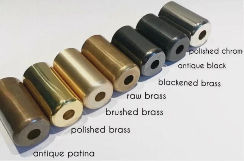 May include: A row of six cylindrical metal hardware pieces with a center hole, labeled as antique patina, polished brass, brushed brass, raw brass, blackened brass, antique black, and polished chrome, arranged diagonally on a white background.