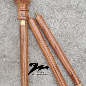 Puede incluir: Bastón de madera con un mango redondeado y un eje desmontable. El bastón es de color marrón claro con detalles dorados. El logotipo "DECOR INTERNATIONAL" es visible en el bastón.