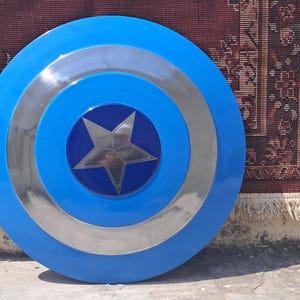 Op de afbeelding: Een blauw en zilver Captain America-schild met een centraal sterembleem. Het schild heeft concentrische cirkels van blauw en zilver. Een klassiek superheldenaccessoire.