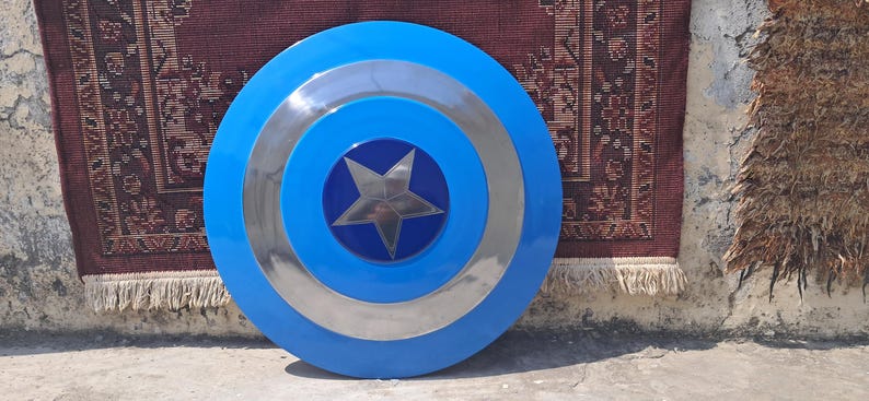 Op de afbeelding: Een blauw en zilver Captain America-schild met een centraal sterembleem. Het schild heeft concentrische cirkels van blauw en zilver. Een klassiek superheldenaccessoire.