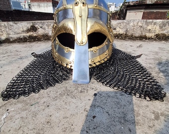 May include: A Viking helmet with a silver and gold finish, featuring a face guard and chainmail. The helmet has a polished appearance and is designed with intricate details. The chainmail extends from the base of the helmet.