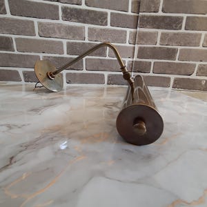 May include: A brass-toned metal picture light featuring a circular wall-mounting plate, a slender adjustable arm with a central hinge, and a cylindrical lamp head with a flat end cap. The fixture has a metallic finish and exposed wiring.