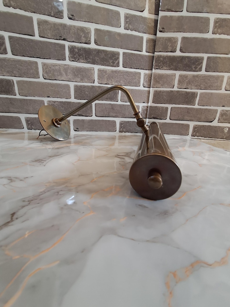 May include: A brass-toned metal picture light featuring a circular wall-mounting plate, a slender adjustable arm with a central hinge, and a cylindrical lamp head with a flat end cap. The fixture has a metallic finish and exposed wiring.